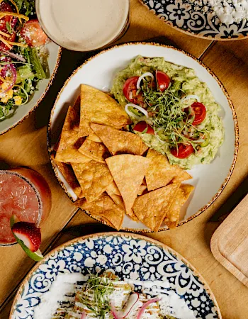 A spread of tortilla chips with guacamole, fresh salads, and creamy dips on wooden boards with limes and herbs.