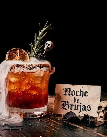A spooky Halloween beer: a frothy mug with smoke, rosemary, lemon, skull decor, and a sign reading “Noche de Brujas.”